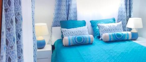 A bedroom with a turquoise bedspread, two pillows, and a nightstand with a lamp.