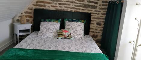 A bedroom with a stone wall, a bed with floral bedding, a nightstand, and a green curtain.