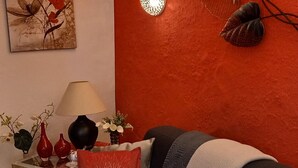 Een moderne woonkamer met een rode accentmuur, een zwarte bank met rode en grijze kussens, een bijzettafeltje met een lamp en decoratieve items, en een kunstwerk in een lijst aan de muur.