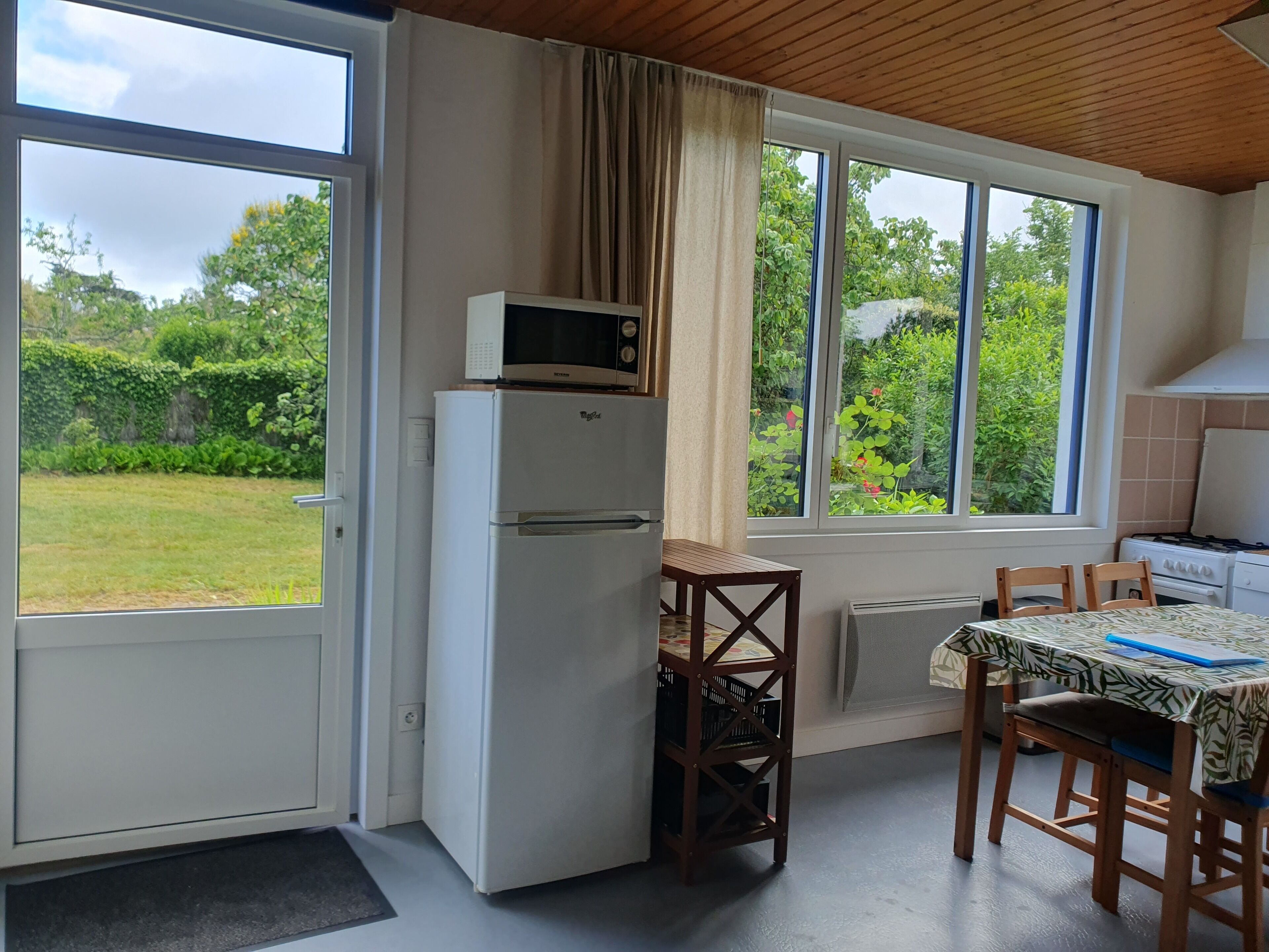 A compact kitchen with a refrigerator, microwave, and stove. There is a dining table with chairs and a view of a garden through large windows.