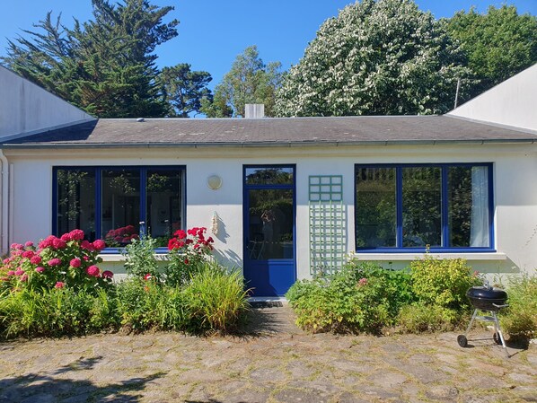 A small house with a blue door, large windows, and a garden with red flowers.