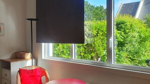 Een kamer met een groot raam, een rode tafel, rieten stoelen, een zwart scherm en een radiator.