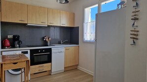 A compact kitchen with wooden cabinets, a built-in oven, a dishwasher, and a window with a view. - The king of grau boucanet quiet apartment in villa entirely renovated (Le Grau-Du-Roi)