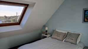 A bedroom with a sloped ceiling, a bed with white bedding, two bedside tables, a nightstand, a lamp, and a window with a view of greenery. - Seaside house in South Finistère, 300m from the beach. (Combrit)