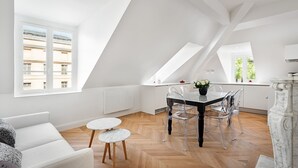 Un loft moderno con comedor, que incluye una mesa negra y sillas transparentes, un sofá blanco y piso de madera con patrón de espiga.