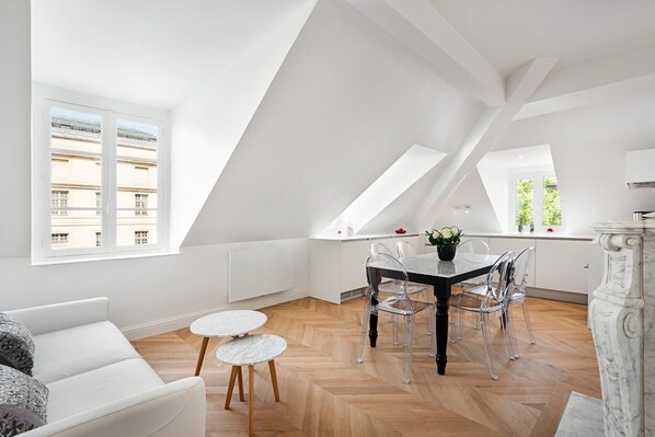 Un appartement moderne de style « loft » comprenant un coin repas avec une table noire et des chaises transparentes, un canapé blanc et un parquet en bois disposé en chevrons.