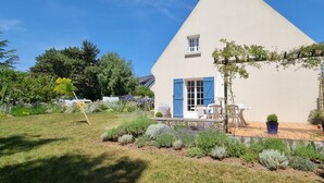 Una casa con techo triangular, un porche pequeño con sillas y un jardín con diversas plantas.