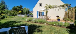 Una casa de fachada blanca, con contraventanas azules y un jardín con diversas plantas y un patio amoblado.