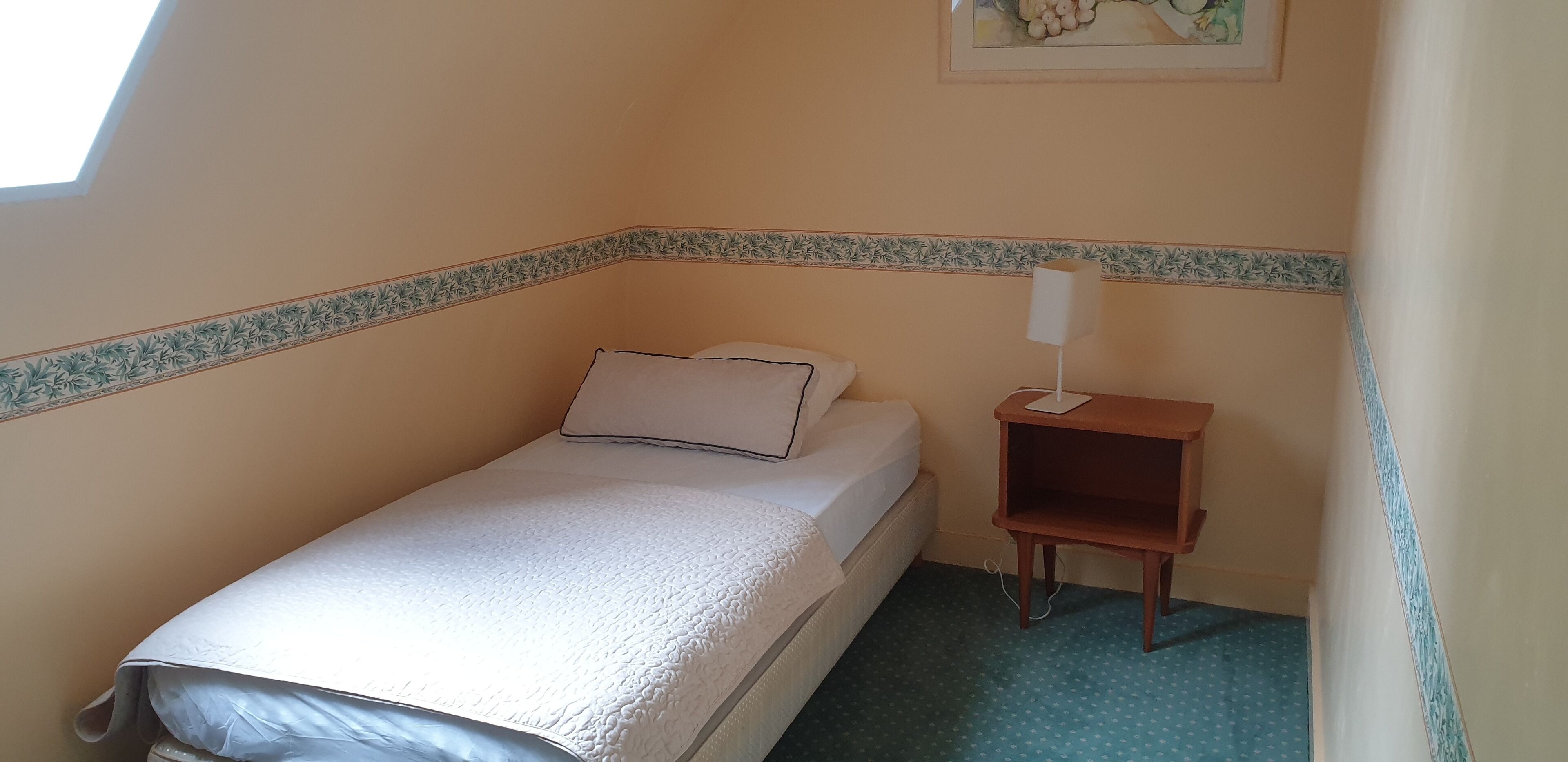 A small, single-bed room with a sloped ceiling, a bedside table with a lamp, and a wall-mounted picture.