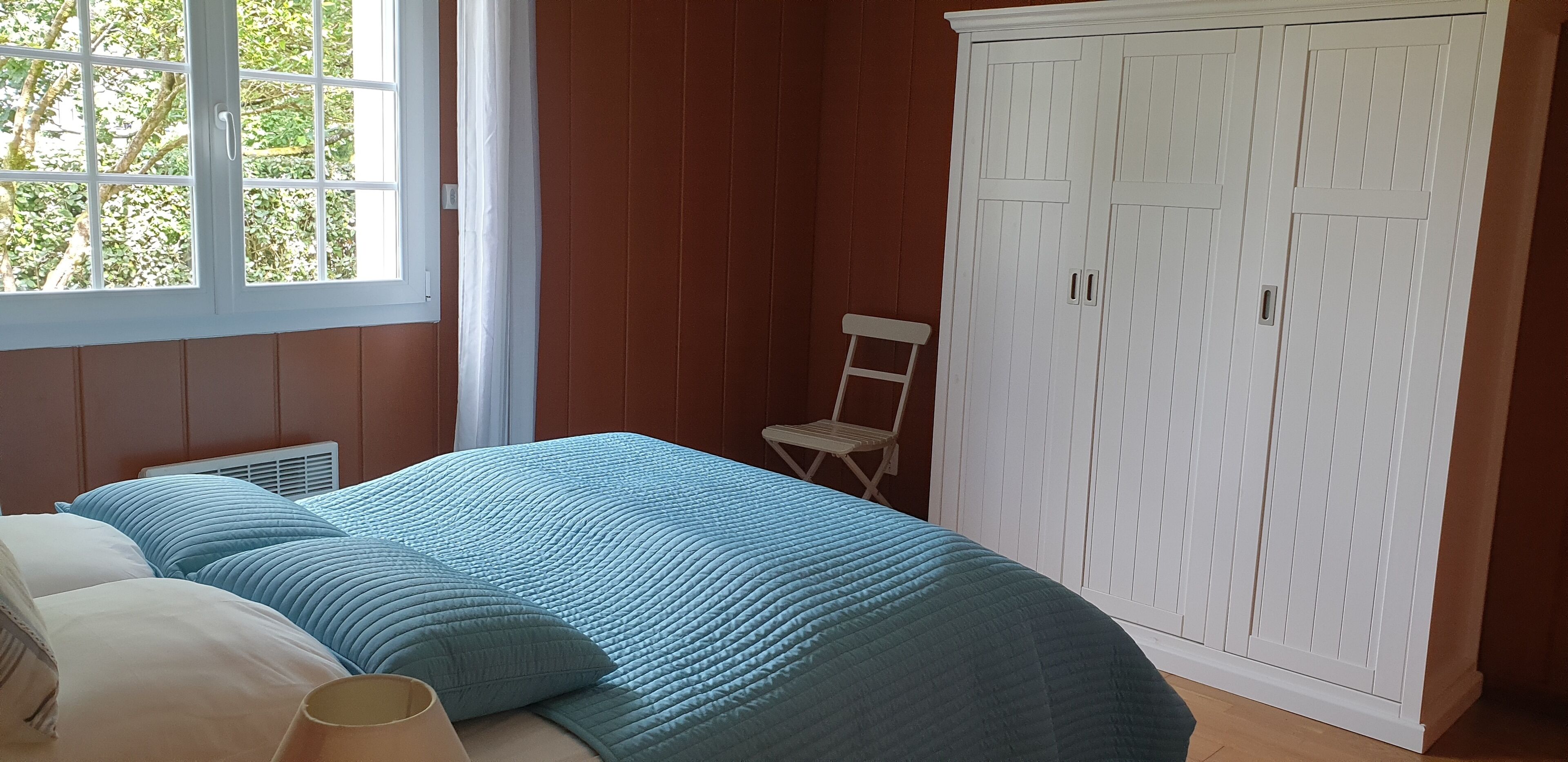 Een slaapkamer met een bed, een raam, een witte kledingkast en een stoel.
