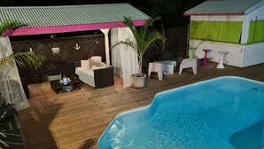 Una piscina con muelle de madera, un área de descanso techada con mobiliario blanco y una pequeña mesa con una planta.
