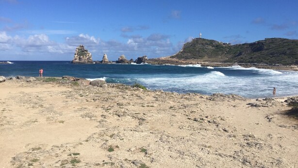 Una playa con formaciones rocosas y una colina con una cruz.