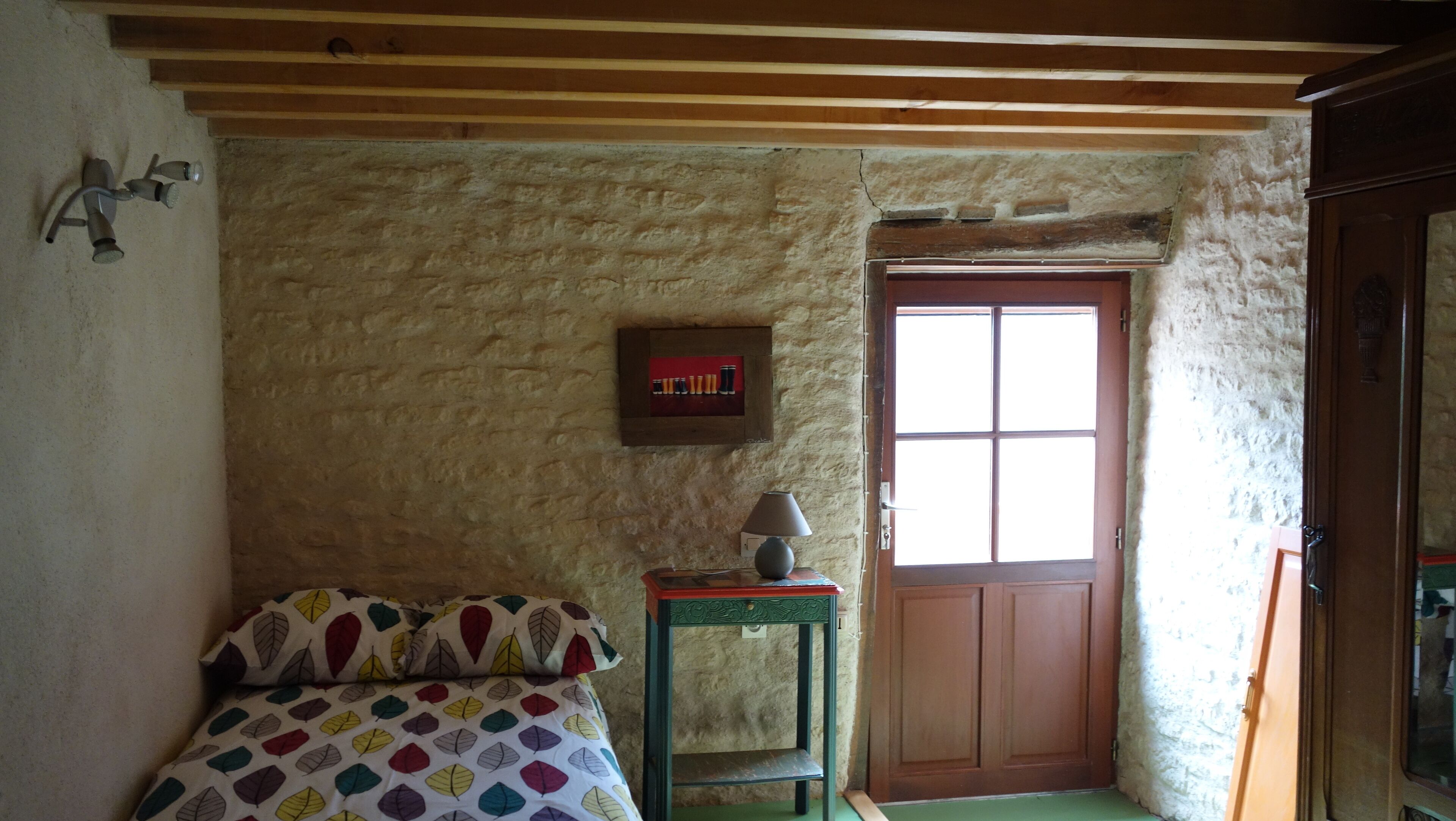 Een eenvoudige kamer met een houten plafond, een bed met kleurrijk beddengoed, een klein tafeltje met een lamp, een deur met een raam en een aan de muur hangend fotolijstje.
