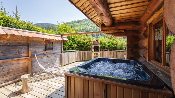 A wooden deck with a hot tub, wooden cabin, and a view of a forested mountain. - La fuste des Chevaux, log cabin, kota-sauna, Nordic bath, 6 people (La Bresse)