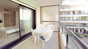 Un balcón con una mesa y sillas blancas, una puerta corrediza de vidrio que da a un dormitorio y vistas de un edificio de varios pisos.