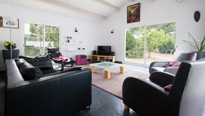 Ein modernes Wohnzimmer mit einer Couch, Sesseln, einem Couchtisch und einem Fernseher. Große Fenster und eine Schiebetür führen in einen Außenbereich.