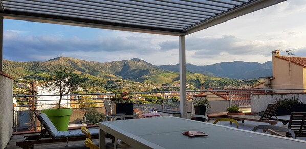 A covered outdoor area with a table and chairs, a green planter, and a view of mountains and a town.