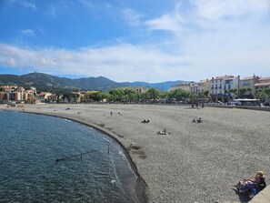 A pebbled beach with people sunbathing and walking, buildings in the background, and mountains in the distance.