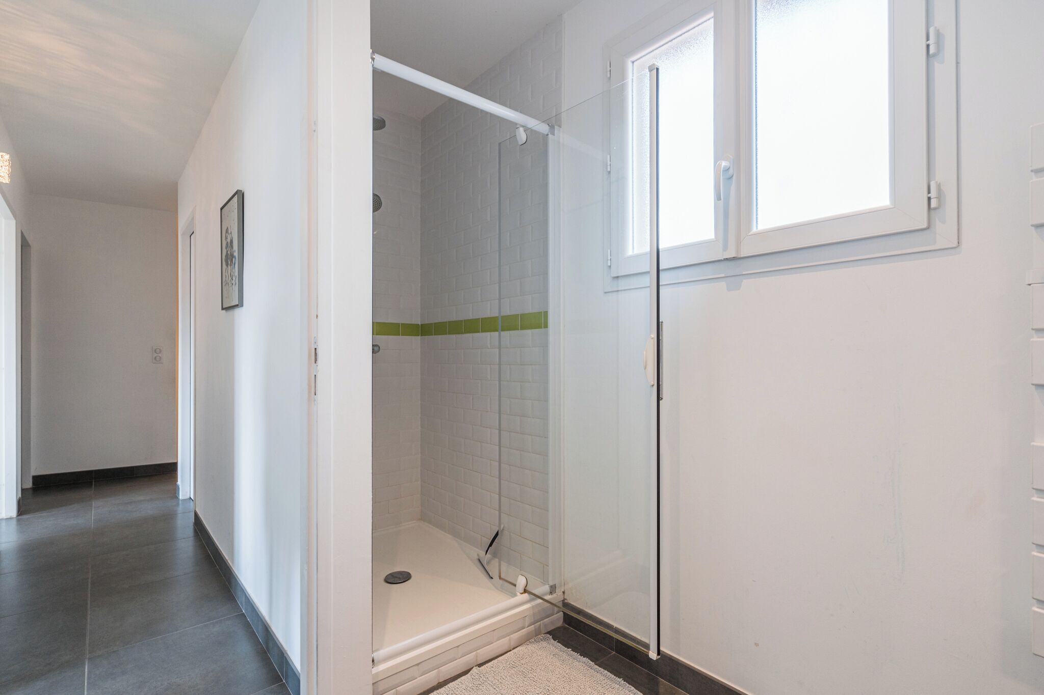 A bathroom with a glass shower enclosure, white tiled walls, and a window.