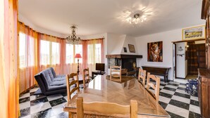 Uma sala de jantar com uma grande mesa de vidro, rodeada por cadeiras de madeira, um sofá e uma lareira.