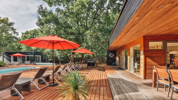 Een houten terras met loungesessels en rode parasols, een zwembad en een modern gebouw met grote ramen.