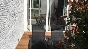 Ein moderner Außenbereich mit Holzterrasse, einer Glastür zu einer überdachten Terrasse und Blick auf ein Wohngebäude.