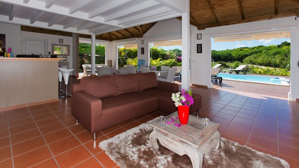 Un espacio habitable amplio con un sofá marrón, una mesa de centro y un jarrón rojo con flores. Se ve una piscina a través de la puerta abierta.