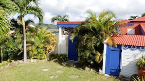 A tropical garden with palm trees, a blue gate, and a red-roofed building. - Sainte-Anne, typical villa for 4-6 people Sea view, directly on the beach. (Sainte Anne)