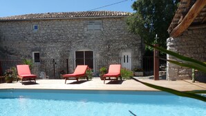 A stone building with a pool, three red lounge chairs, and a wooden deck.