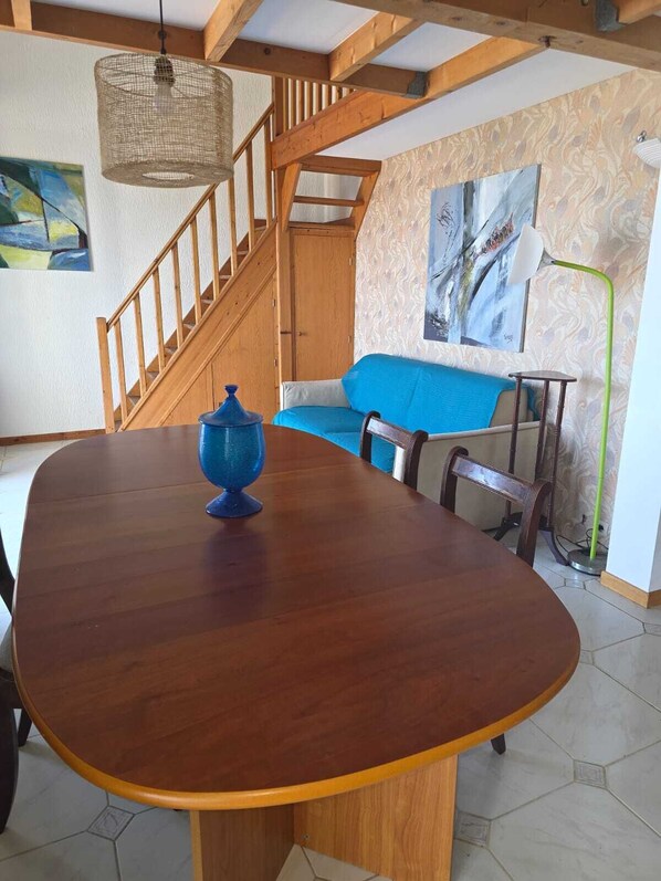 A dining area with a wooden table, a blue decorative item, a staircase, a blue sofa, and a painting on the wall.