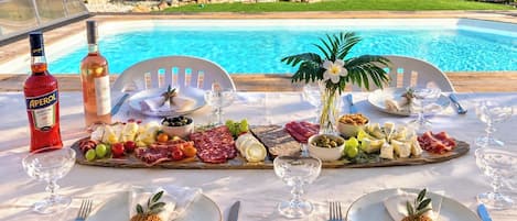 A table set for a meal with a pool view, featuring a variety of cheeses, meats, and fruits.