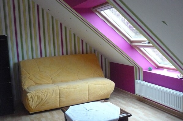 Une pièce avec un canapé jaune, une petite table, une fenêtre donnant sur un paysage et un radiateur.