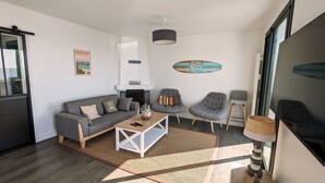Een moderne woonkamer met een grijze bank, een bijzettafeltje en twee fauteuils. Er is een open haard, een hanglamp en een groot raam.