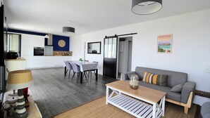Een moderne woonkamer met een grijze bank, een houten salontafel en een eethoek met een witte tafel en stoelen.
