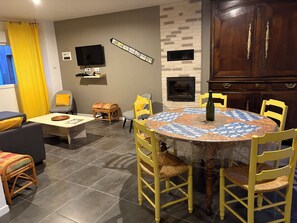 Un salon avec une table ronde, des chaises jaunes, un téléviseur à écran plat, une cheminée et un rideau jaune.