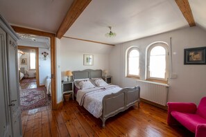 Ein Schlafzimmer mit Holzboden, einem Bett mit weißem und grauem Kopfteil, zwei Nachttischen mit Lampen, einem roten Sessel und einem Fenster mit Blick ins Freie.
