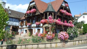 Une maison traditionnelle à colombages, dotée d’un toit à forte pente, de plusieurs balcons ornés de plantes en fleurs et d’une cheminée bien visible.