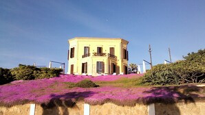 Un bâtiment à deux étages doté d’un balcon au deuxième étage, situé sur une colline et agrémenté de fleurs violettes.
