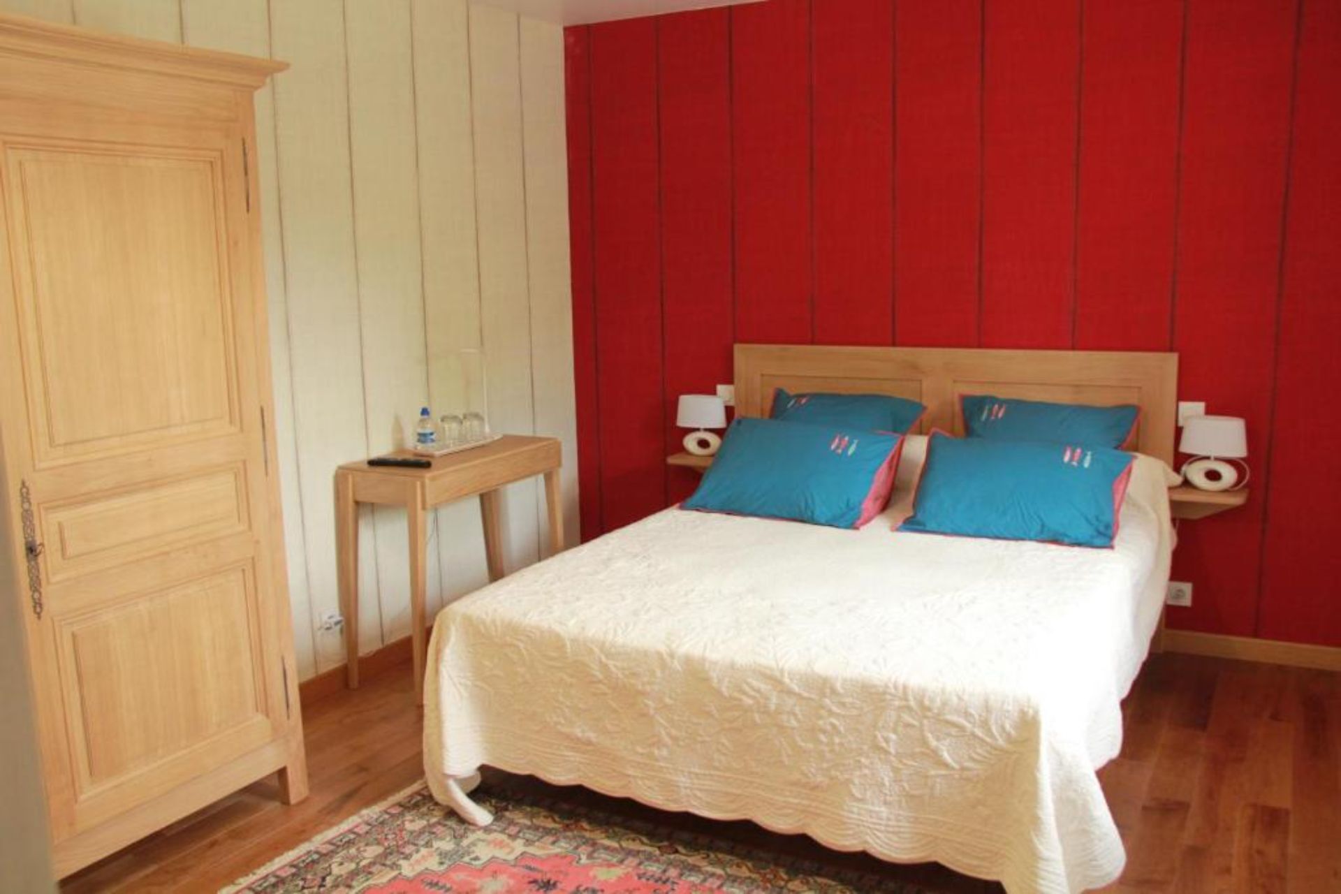 A bedroom with a wooden wardrobe, a bedside table with a lamp, and a bed with blue pillows.