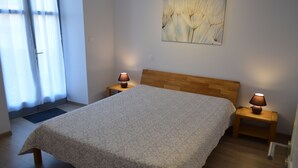 A bedroom with a wooden headboard, a large bed with a floral bedspread, two bedside tables with lamps, a window with sheer curtains, and a wall-mounted artwork.