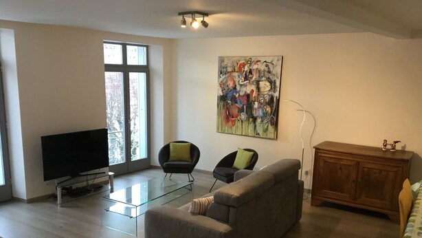 A modern living room with a grey sofa, a black armchair, a glass coffee table, a flat-screen TV, and a wooden cabinet.