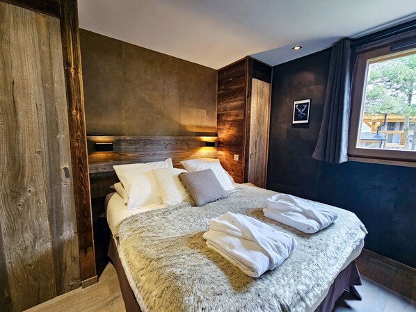 A bedroom with a wooden headboard, a large bed with white linens, a fluffy blanket, and a window with a view of trees. - Chalet Kasumiga Collection Perles (Bolquère)