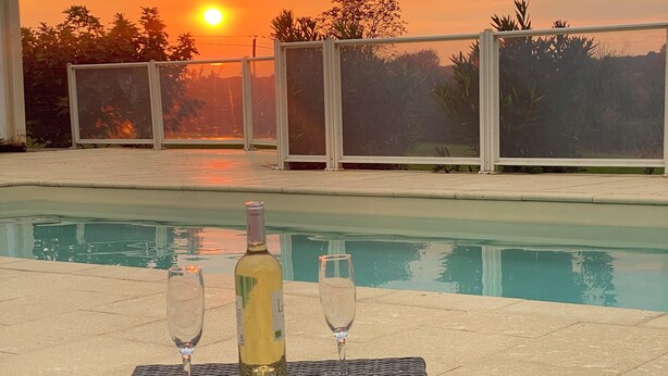 Une table au bord de la piscine, avec deux verres à vin et une bouteille, le tout mis en valeur par un magnifique coucher de soleil en arrière-plan.