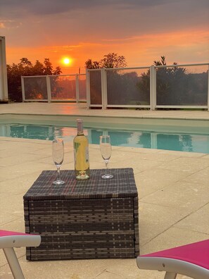 A poolside table with two wine glasses and a bottle, set against a sunset view.