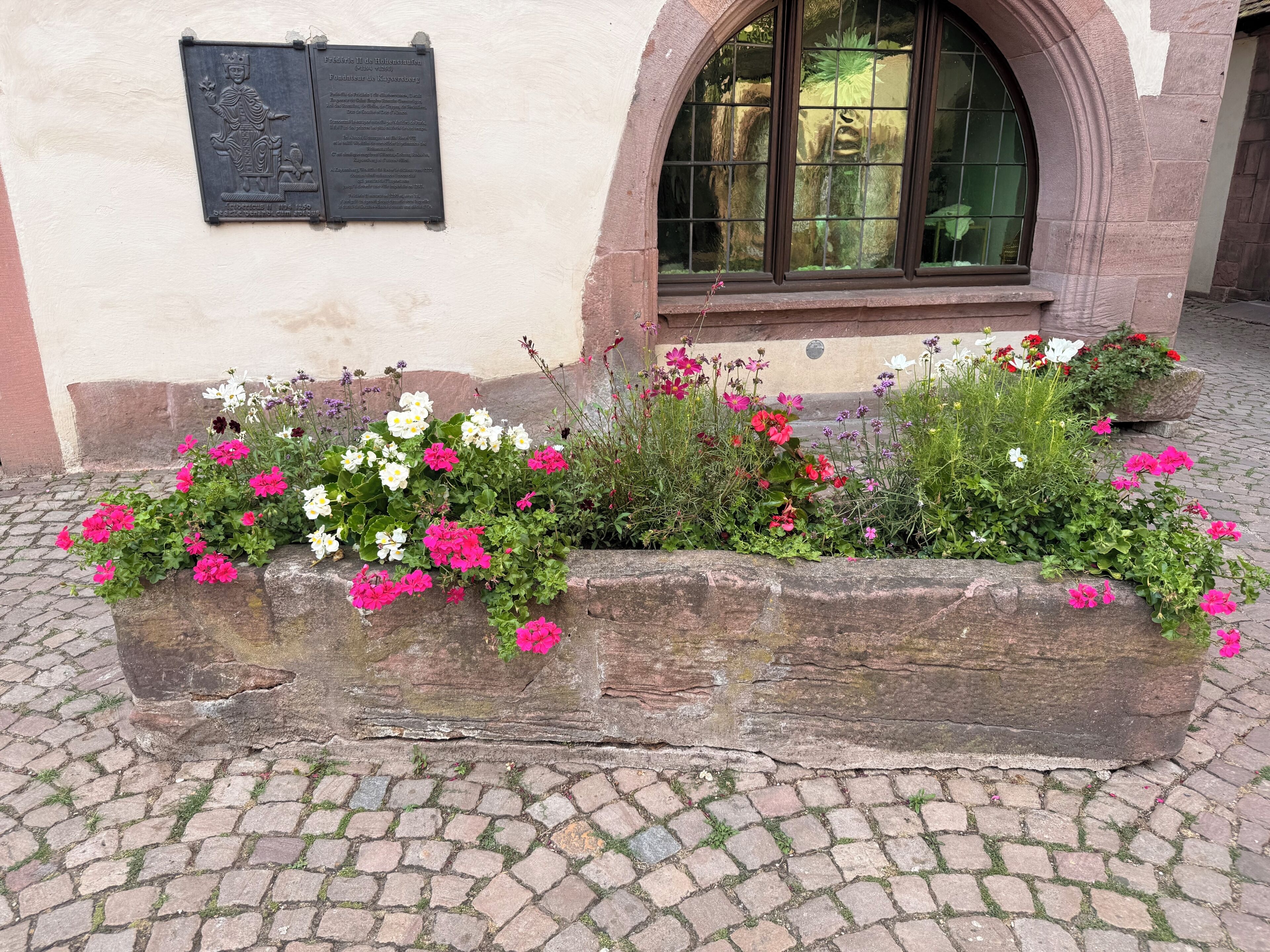 Un bac à fleurs en pierre, orné de fleurs, se trouve devant un bâtiment sur lequel on peut voir une plaque commémorative et une fenêtre en arc de cercle.