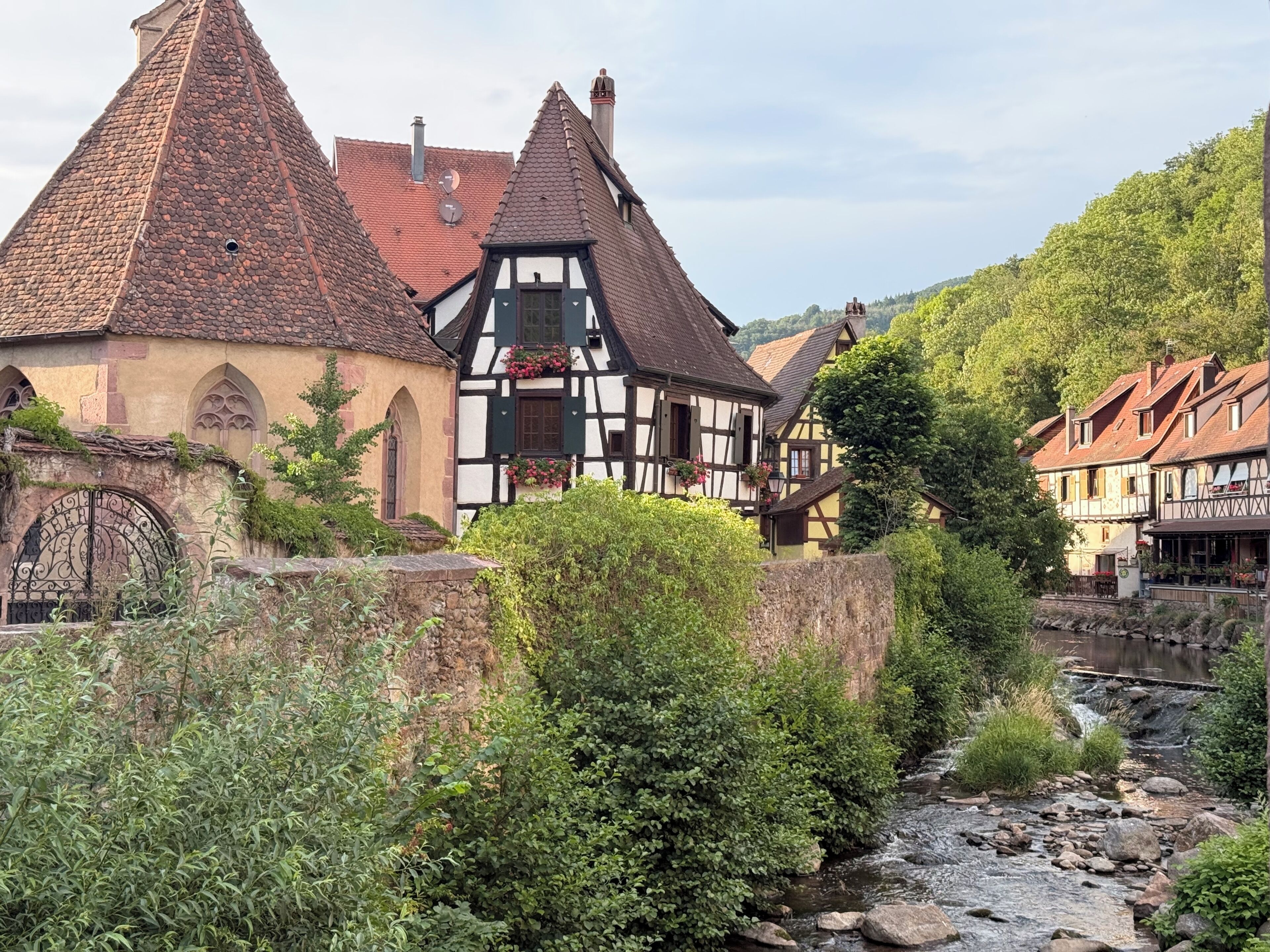 Traditional half-timbered houses with steeply pitched roofs, a stone wall, and a stream running through a lush green landscape.