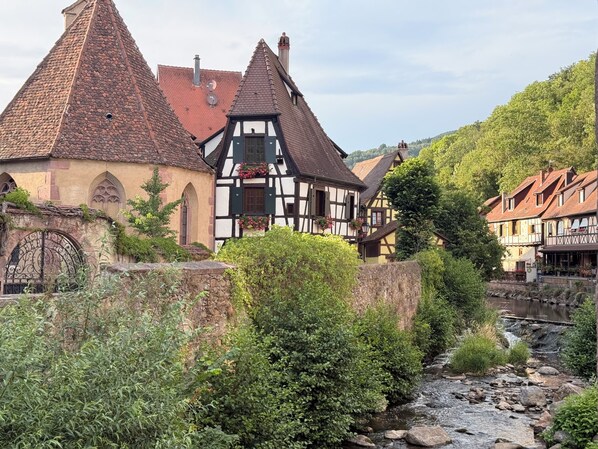 Traditional half-timbered houses with steeply pitched roofs, a stone wall, and a stream running through a lush green landscape. - New flat in a typical village near Colmar and alsatian vineyard (Wickerschwihr)