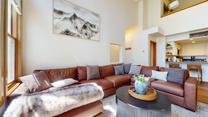 A modern living room with a large brown sectional sofa, a round coffee table, and a mountain landscape painting on the wall.