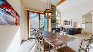 A spacious dining area with a wooden table and chairs, a large window with a view of trees, and a modern chandelier. - Snowcreek #880 3 Bedroom Condo by RedAwning (Mammoth Lakes)