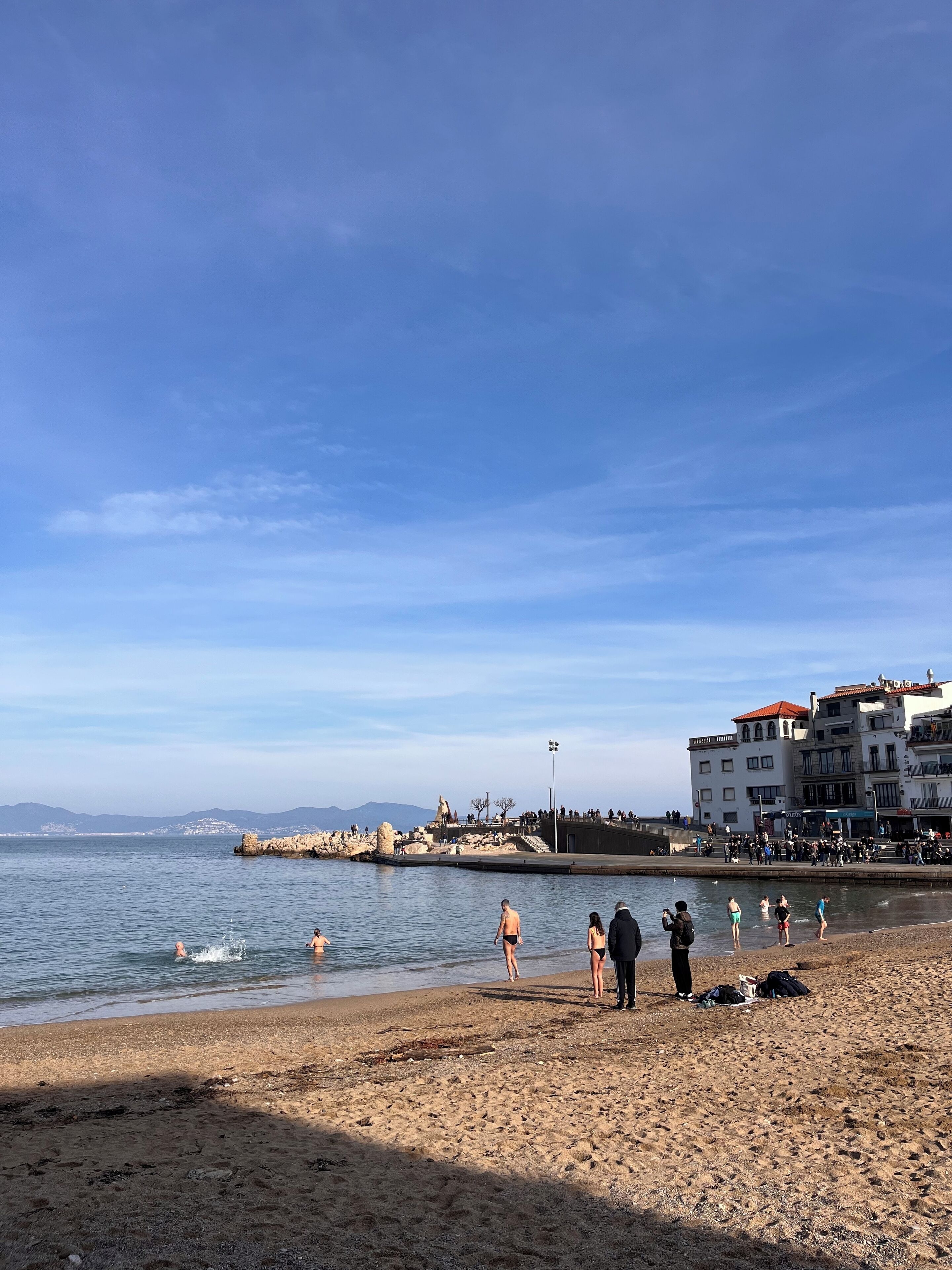 A beach with people swimming and walking along the shore, buildings in the background, and a clear sky.